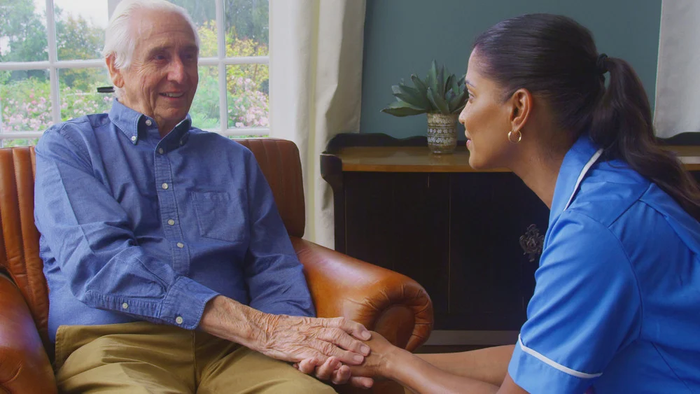 Senior man holding caregiver's hands while both are seated in a cozy living room.
